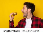 Closeup Side View Profile Portrait Of Funny Hungry Guy Biting Burger Eating Junk Food Holding Sandwich With Open Mouth Posing On Yellow Studio Background. Man Enjoying Big Hamburger, Bad Eating Habit