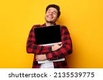 Small photo of Technology Lover Concept. Happy man hugging laptop with black blank empty screen, holding it tight near chest. Casual male geek with closed eyes standing isolated on yellow orange studio wall