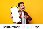 Small photo of Cheerful Emotional Man Holding Big Blank Cell Phone In Hand Showing White Screen To Camera Pointing At You, Happy Millennial Guy Recommending New Application Or Mobile Website, Mockup Banner Collage