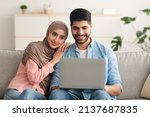 Middle Eastern Husband And Wife Using Laptop Watching Movie Online Embracing Sitting On Couch At Home. Front View Shot Of Arabic Spouses Browsing Internet Via Computer Indoor. Technology Concept
