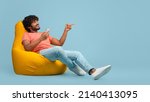 Happy curly millennial indian man sitting at bean bag with brand new smartphone and pointing at copy space over blue studio background, showing nice offer and smiling, panorama