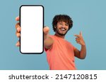 Small photo of Cool handsome curly young indian guy in stylish pink t-shirt showing brand new cell phone with white empty screen and gesturing, sharing exciting online deal, blue background, mockup