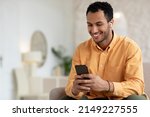 Smiling Arabic Male Listening To Music On Mobile Phone Wearing Wireless Headphones Sitting On Couch Relaxing At Home. Male Enjoying Favorite Playlist Using Musical Application, Free Copy Space