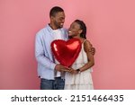 St. Valentines Day Concept. Portrait of happy black couple in love holding red heart shaped balloon, smiling guy embracing his lady, looking at each other, isolated on pink studio background wall