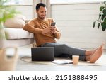 Portrait of young man holding using cell phone sitting on floor leaning on couch in living room. Cool guy browsing internet, surfing web watching video, using application and pc, full body length