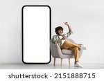 Small photo of Joyful bearded curly indian guy in casual sitting in armchair with brand new smartphone and wireless headset, watching movie or listening to music online while chilling at home, copy space, mockup