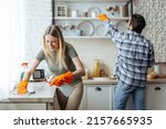Smiling millennial european woman blonde with rubber gloves and man wipe dust on light kitchen interior, copy space. Hygiene, cleaning at home together and household chores during covid-19 outbreak