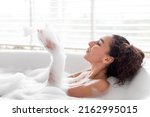 Lovely young female lying in bubble bath, blowing bubble foam from hands at home, copy space. Pretty millennial woman enjoying spa procedure, having fun during domestic body care