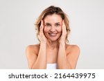 Anti-Aging Skincare. Beautiful Middle Aged Woman Touching Her Face And Smiling At Camera, Attractive Happy Mature Woman Standing Wrapped In Towel Over Light Grey Studio Background, Copy Space