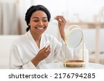 Skincare Concept. Beautiful Black Lady Applying Face Serum With Dropper While Sitting In Front Of Mirror At Home, Smiling African American Woman Moisturizing Skin, Enjoying Self-Care Routine