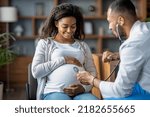 Small photo of Pregnant millennial black woman sitting on chair at clinic, touching her big tummy and smiling, visiting doctor, arab man gynecologist checking baby heart rate with stethoscope, copy space