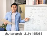 Small photo of English Course. Happy Asian Male Teacher Having Foreign Language Class Standing Pointing At Whiteboard With Grammar Rules Smiling To Camera Posing In Modern Classroom At School. Education Concept