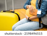 Small photo of Giving birth abroad concept. Unrecognizable pregnant woman travelling alone, sitting on bench with backpack next to baggage, holding passport, flight tickets, hugging tummy, copy space, cropped