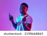 Small photo of Portrait of cheerful young black man holding cellphone and credit card, using banking or shopping mobile app, receiving cashback, trading or gambling online in neon light