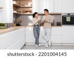 Small photo of Cheerful asian couple enjoying morning coffee at home, happy pretty young lady having conversation with her husband, standing by kitchen table, holding mugs and chatting, full length, copy space