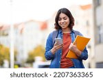 Small photo of Smiling Young Middle Eastern Female Student Walking With Smartphone And Workbooks On City Street, Happy Millennial Arab Woman Messaging On Mobile Phone While Going Home After Classes, Copy Space