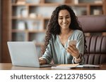 Happy attractive curly middle eastern young woman sitting at workdesk with laptop and notepads on, using smartphone and smiling, using newest business mobile app, office interior, copy space