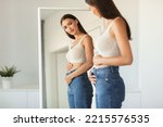 Young Woman Touching Flat Belly Expecting Baby And Enjoying First Months Of Pregnancy Posing Looking At Her Reflection In Mirror Wearing Skinny Jeans Standing At Home. Selective Focus