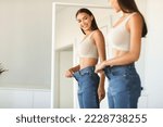 Small photo of Excited young fit woman losing weight and wearing old too big jeans, woman feeling satisfied with results of her diet and slimming, posing near mirror, free space