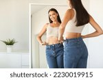 Cheerful young woman posing wearing jeans and smiling to her reflection in mirror after successful weight loss, copy space. Female beauty and style, self confidence, slimming concept