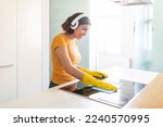 Happy Middle Eastern Female Listening Music In Headphones While Cleaning Kitchen, Cheerful Young Arab Woman Polishing Induction Cooktop Surface With Rag And Enjoying Favorite Songs, Copy Space