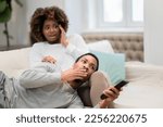 Small photo of Scared shocked millennial african american man and woman couple spouses sitting on couch, gesturing, watching appalling content on TV, copy space for advertisement
