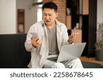 Small photo of Confused korean man looking at cellphone screen, sitting on couch with computer on lap, frustrated male reading bad news, having problem with device or Internet connection