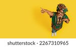 Small photo of Cheerful happy pretty young black woman in traditional outwear turban and accessories pointing at copy space, showing advertisement over yellow studio background, high angle view, banner