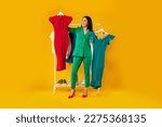 Shopaholic. Happy elegant woman choosing dresses holding two hangers, standing near clothing rail with trendy clothes over yellow background. Female fashion choice