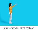 Side view of slim beautiful long-haired young asian woman in casual outfit pointing at free space for advertisement or text, isolated on blue studio background, full length shot