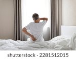 Small photo of Unrecognizable mature grey-haired man wearing white pajamas sittting on bed at home in the morning, touching lower back and neck, suffering from muscle strain after waking up, copy space