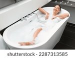 Full Length Shot Of Female Taking Bath Relaxing Lying In Water With Foam At Home. Happy Lady Closing Eyes Enjoying Hygiene Routine In Modern Bathroom Indoor. Selective Focus