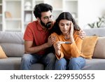 Infertility Problems. Portrait Of Upset Young Indian Couple Looking At Negative Pregnancy Test While Sitting Together On Couch At Home, Caring Eastern Husband Comforting Depressed Wife, Free Space