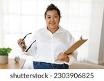 Small photo of Portrait of professional family counselor cheerful attractive young hispanic woman plus size wearing formal outwear posing in clinic cozy office, holding folder and eyeglasses, copy space