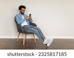 Small photo of Mobile Communication. Young Indian Guy Messaging On Smartphone While Relaxing In Armchair At Home, Happy Handsome Eastern Male Texting With Friends Or Browsing Social Media, Copy Space