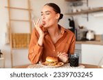 Contented caucasian lady licking her fingers while eating delicious french fries and hamburger, enjoying junk food at home in kitchen interior, copy space