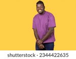 Shy African American young man looking at camera with toothy smile, holding hands together, posing wearing casual purple t-shirt, standing over yellow background, studio shot with free space