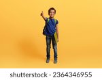 Small photo of Happy cute preteen black schoolboy with backpack and exercisebook showing thumb up and smiling isolated on yellow studio background. Cheerful african american kid going to school, copy space