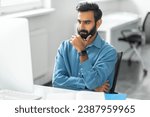 Small photo of Pensive bearded indian man wearing blue shirt and smartwatch, touching chin and looks intently at computer screen in modern, bright office setting