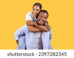 Delighted black couple enjoying piggyback ride, woman laughing and embracing the man from behind, set against bright yellow background