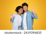 Small photo of Affable happy young African American woman and man waving hello with open hands, both sporting bright smiles and casual blue attire, set against a cheerful yellow backdrop