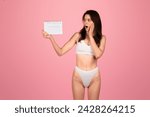 Small photo of Shocked european young woman in white underwear holding a calendar with a surprised expression, symbolizing forgotten dates or events on a pink background, studio. Body care, fit lifestyle