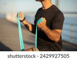 Unrecognizable sporty man doing lunge exercising with resistance rubber band outdoor at seaside setting, cropped shot, showcasing toned muscles and dedication to fitness and sport. Selective focus