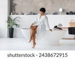 Beautiful young black woman wearing white bathrobe sitting on bathtub and touching her beautiful legs, happy female enjoying silky soft skin after morning shower, posing in modern bathroom interior