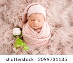Small photo of Cute newborn baby girl sleeping swaddled in fabric and wearing hat. Infant kid studio portrait with flowers during napping