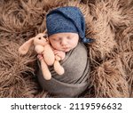 Small photo of Newborn baby boy swaddled in fabric sleeping and holding knitted bunny toy in his hands. Infant kid napping on fur