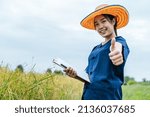 Small photo of Portrait Asian Young farmer woman wearing straw hat and holding clipboard in hand, she smile and show thump up to camera during work on rice field, copy space and selective focus