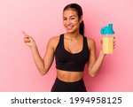 Young Venezuelan woman drinking a protein shake isolated on pink background smiling and pointing aside, showing something at blank space.