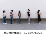 Group of young people at safety distance near a white wall in queue in protection by Coronavirus, Covid-19 with face mask while using the device - Multiracial group of friends - Concept of security