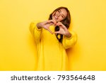 Smiling young girl in yellow sweater showing heart with two hands, love sign. Isolated over yellow background.
